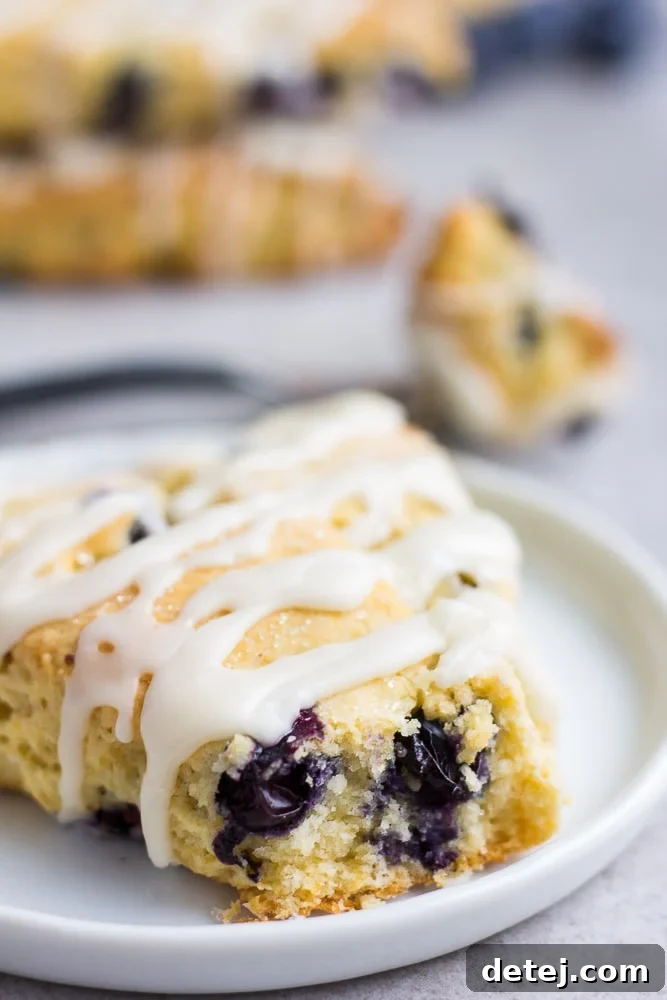 A single glazed vegan blueberry scone elegantly placed on a white plate, with a soft-focus background of more scones.