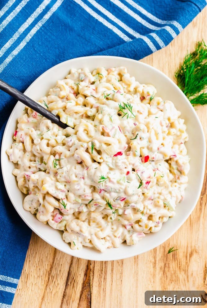 A large bowl brimming with vibrant vegan macaroni salad, accompanied by a serving spoon and a soft blue towel in the background, signaling a refreshing summer dish.