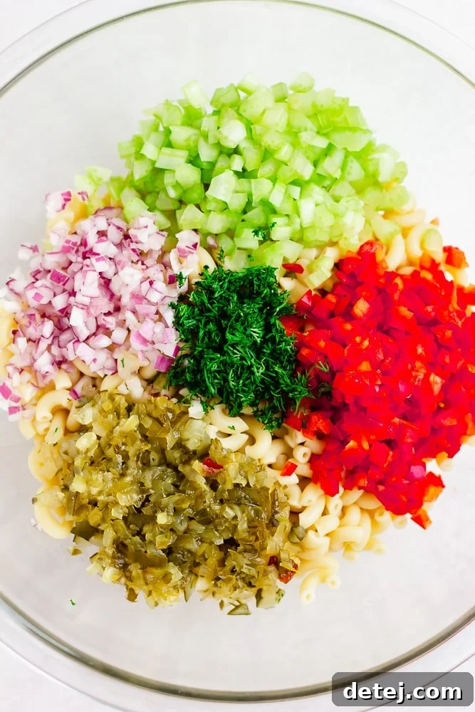 A vibrant assortment of prepared ingredients for vegan macaroni salad, including cooked elbow noodles, finely chopped red bell pepper, red onion, fresh dill, sweet pickles, and crisp celery, all ready to be mixed.