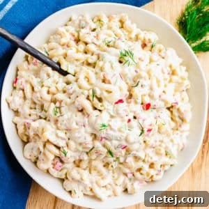 A pristine white bowl overflowing with creamy macaroni salad, set against a blurred background with a blue towel, highlighting its fresh appeal.