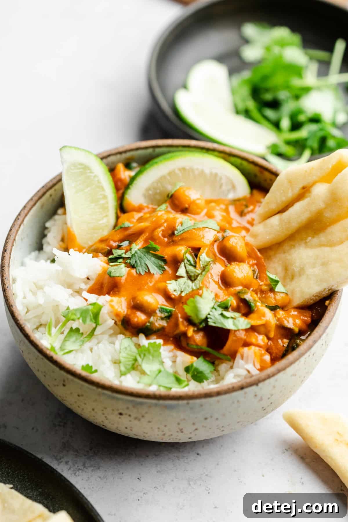 A bowl of chickpea curry served with rice, fresh cilantro, a lime wedge, and a piece of naan bread on a rustic grey background.