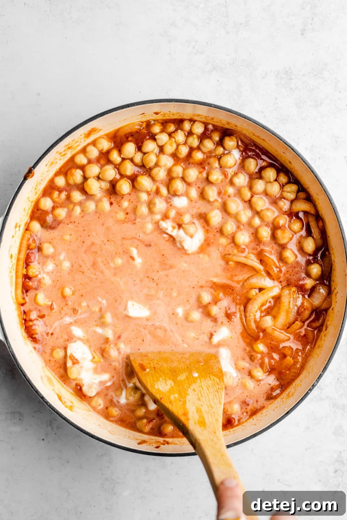 Pouring creamy coconut milk into a pot containing chickpeas and a reddish tomato-based sauce with sautéed onions, enhancing the richness.