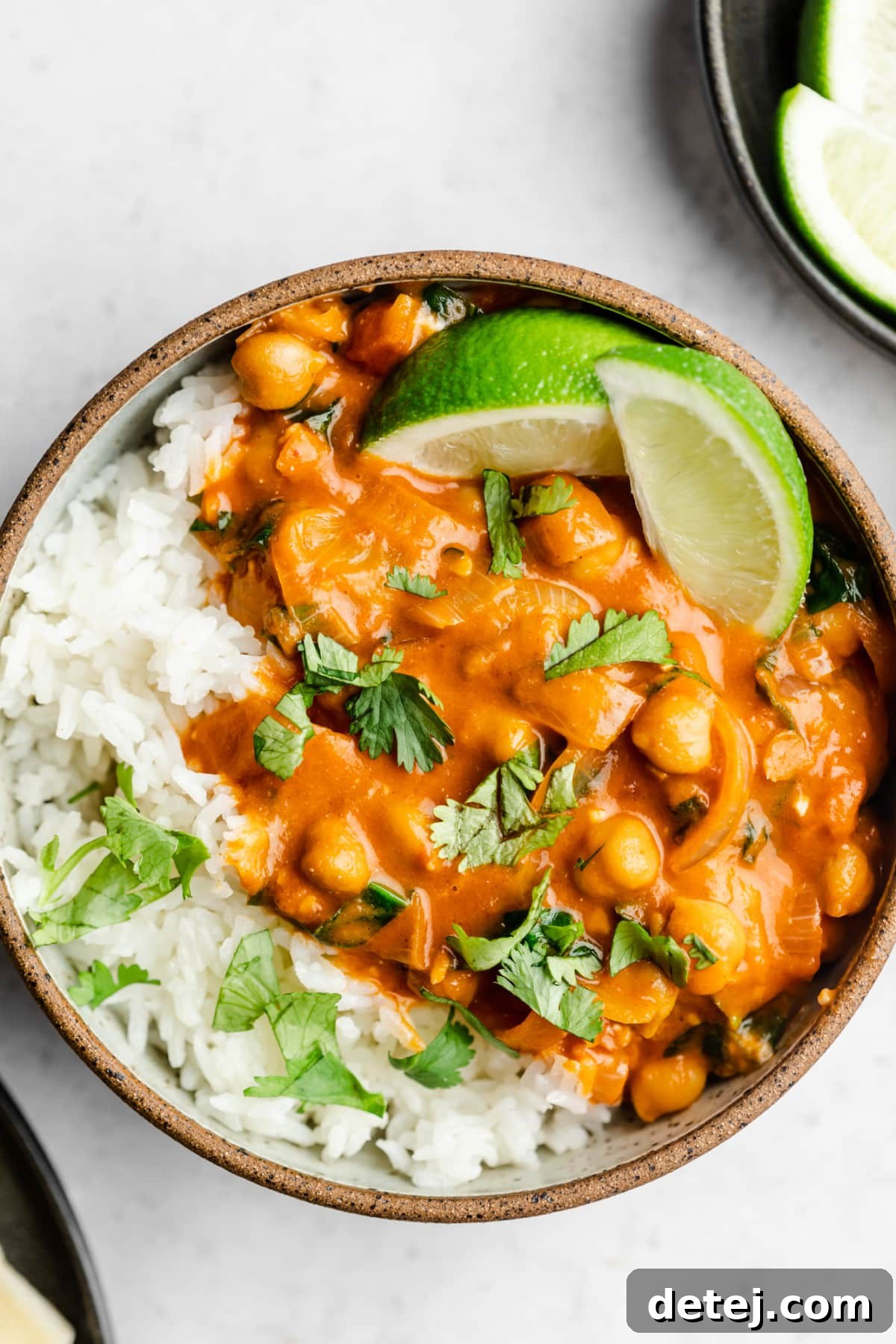 A close-up overhead shot of a bowl filled with creamy reddish chickpea curry, garnished with lime wedges and fresh cilantro, alongside a serving of rice.