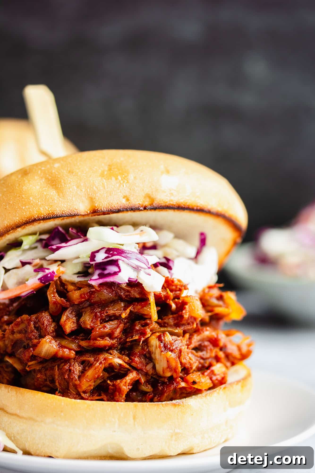 Close up on a BBQ Jackfruit Pulled Pork sandwich topped with creamy coleslaw, served on a white plate. This vegan delight offers a smoky and savory experience that rivals the real thing.