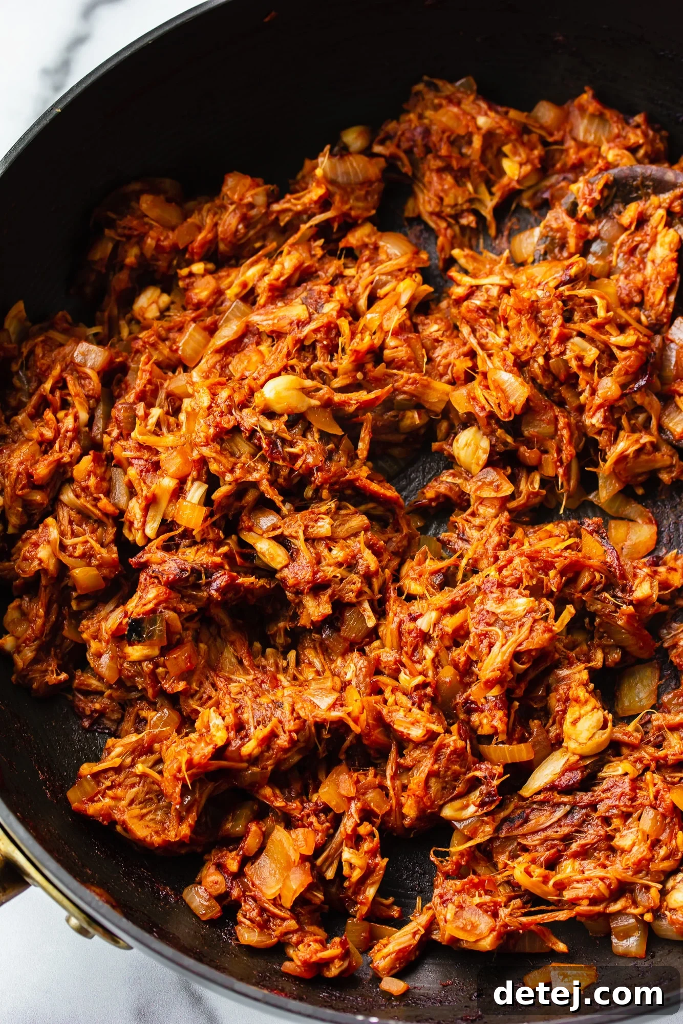 Stirring BBQ jackfruit pulled pork with a wooden spoon in a black skillet, showcasing the rich sauce and shredded texture.