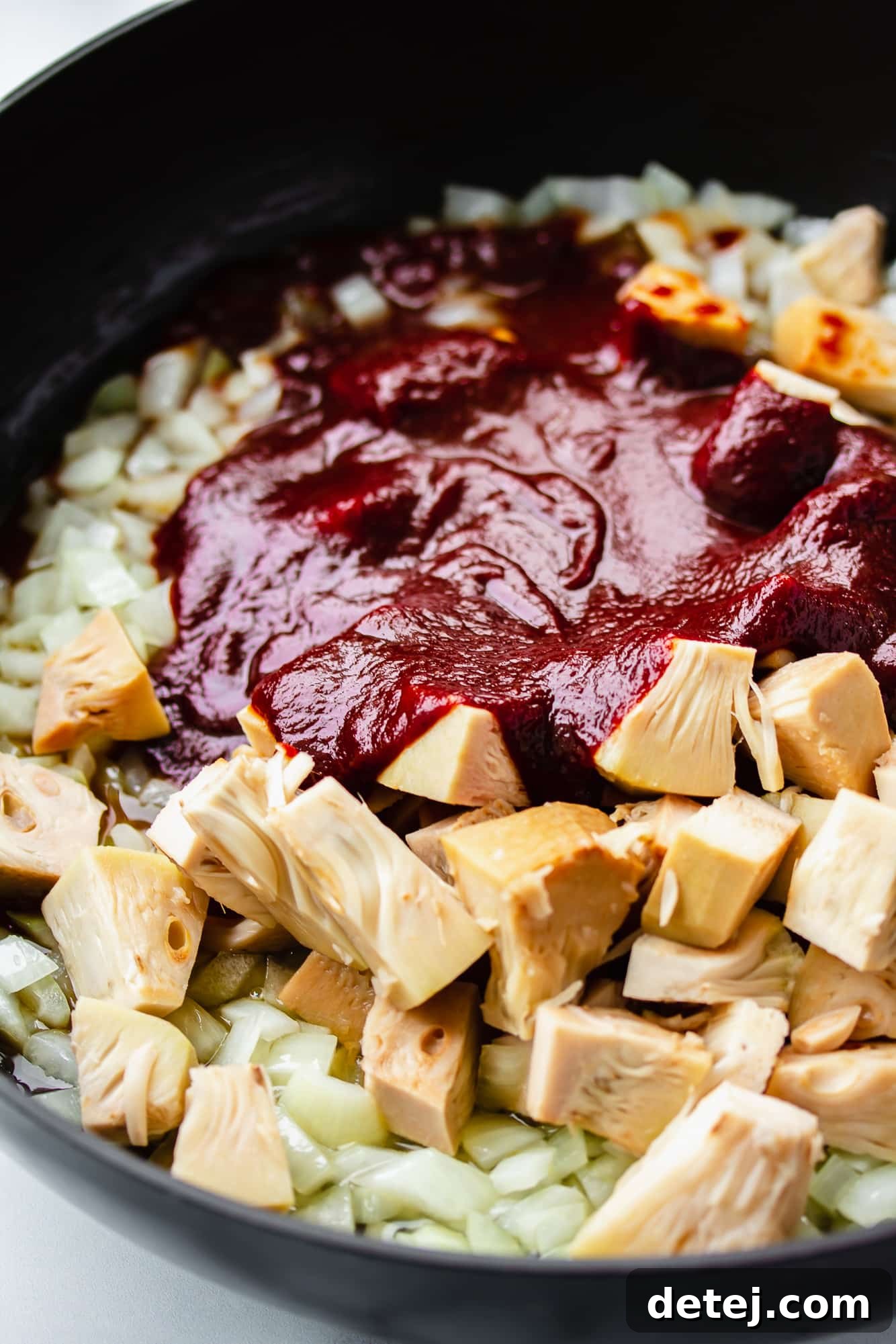 Jackfruit cooking in a large black skillet with diced onion and a rich barbecue sauce, simmering to tender perfection.