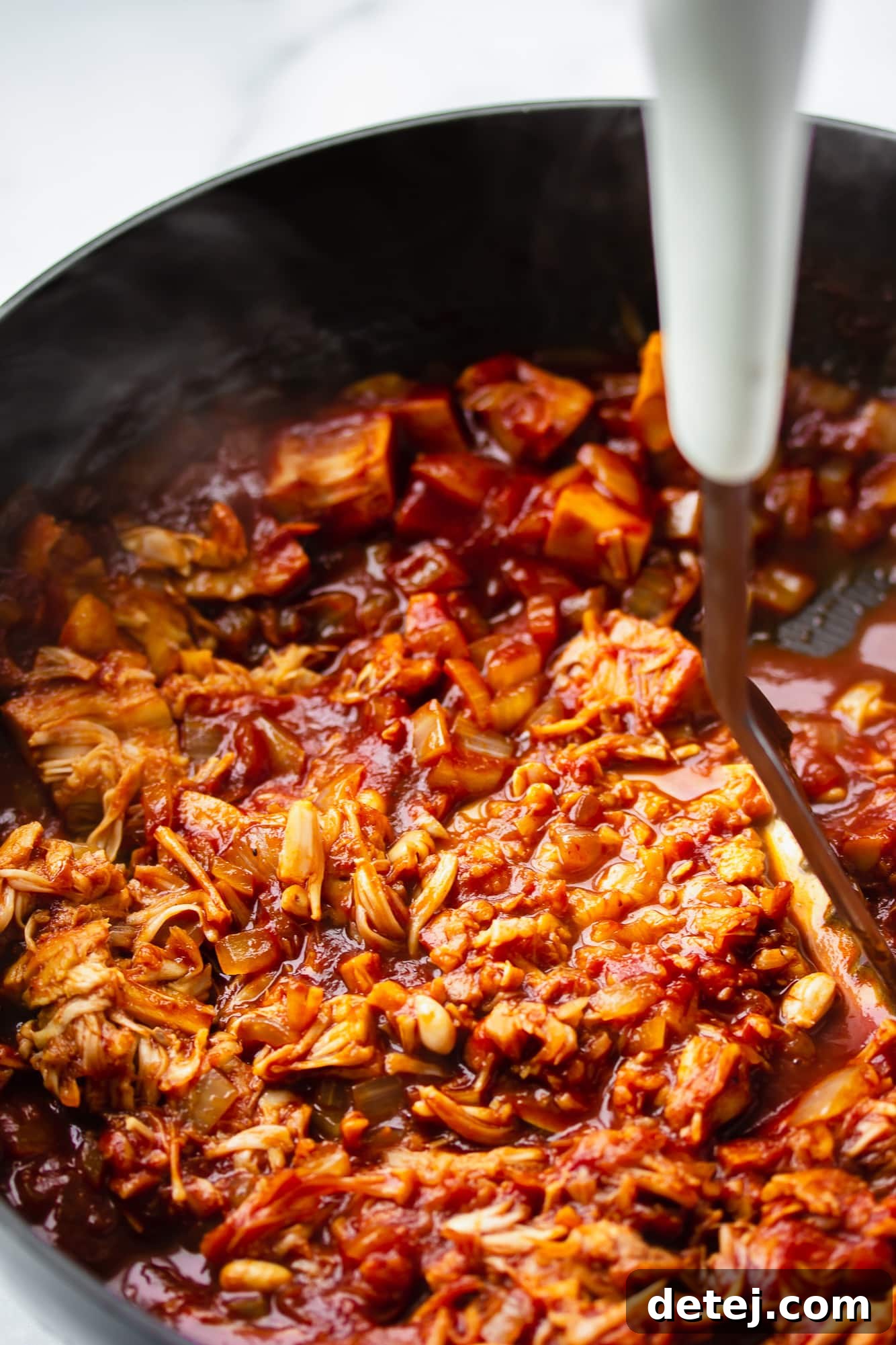 Mashing vegan pulled jackfruit in a skillet with a potato masher to create a shredded, meat-like texture.