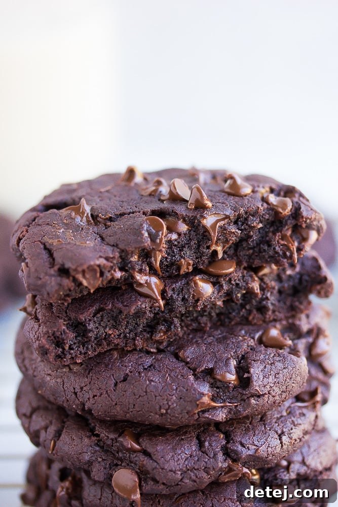 A broken vegan chocolate cookie resting on a stack of whole cookies, showcasing its soft, fudgy interior and abundance of chocolate chips.