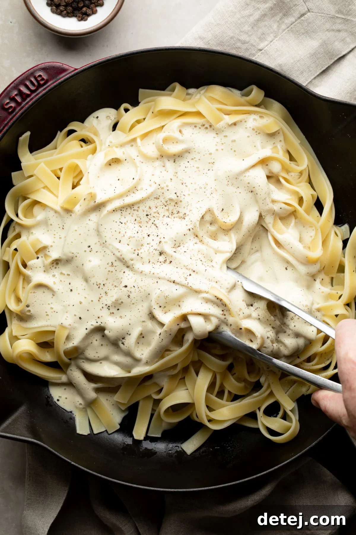 white sauce poured on a cast iron pan with cooked noodles, tongs getting ready to mix