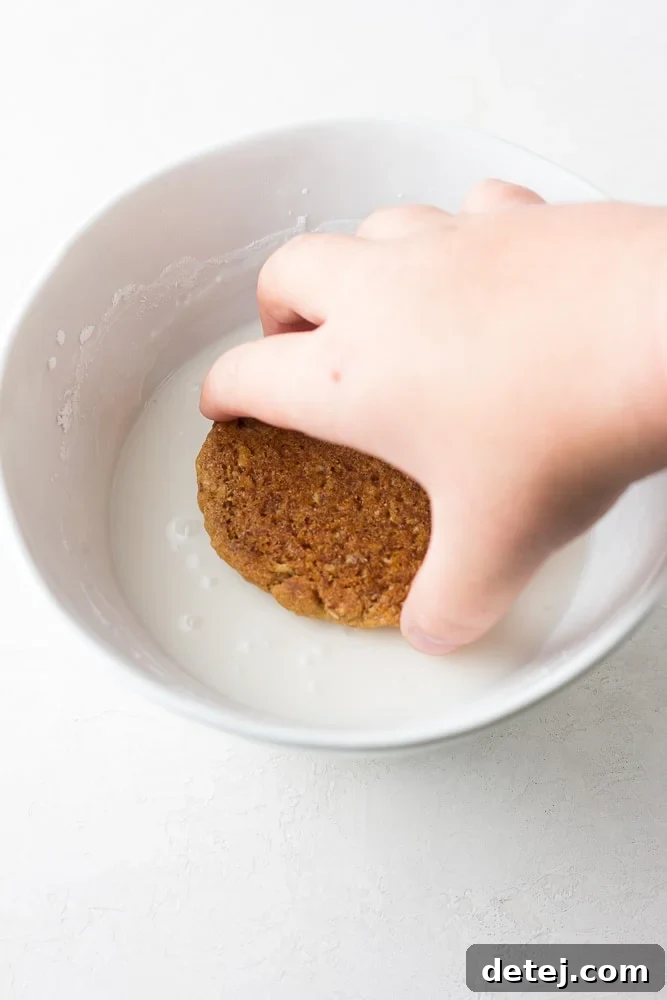 A vegan oatmeal cookie being gently dipped into a bowl of thick white icing, illustrating the icing process.