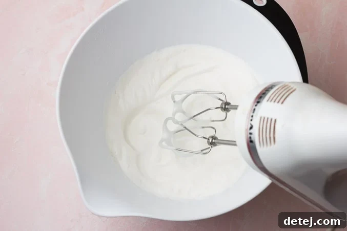 A hand mixer rapidly beating chickpea liquid in a bowl, showing the mixture turning fluffy and increasing in volume.
