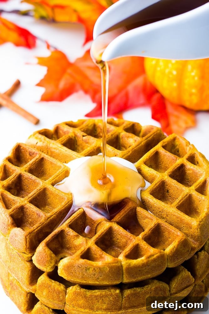 maple syrup pouring over vegan pumpkin waffles, with autumn leaves and mini pumpkins in the background