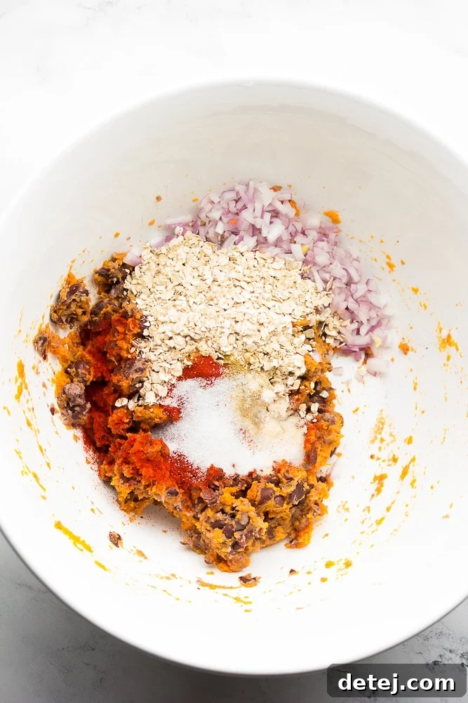 White bowl filled with unmixed ingredients for the sweet potato black bean burgers: mashed sweet potato and black beans, diced red onion, oats, and various spices.