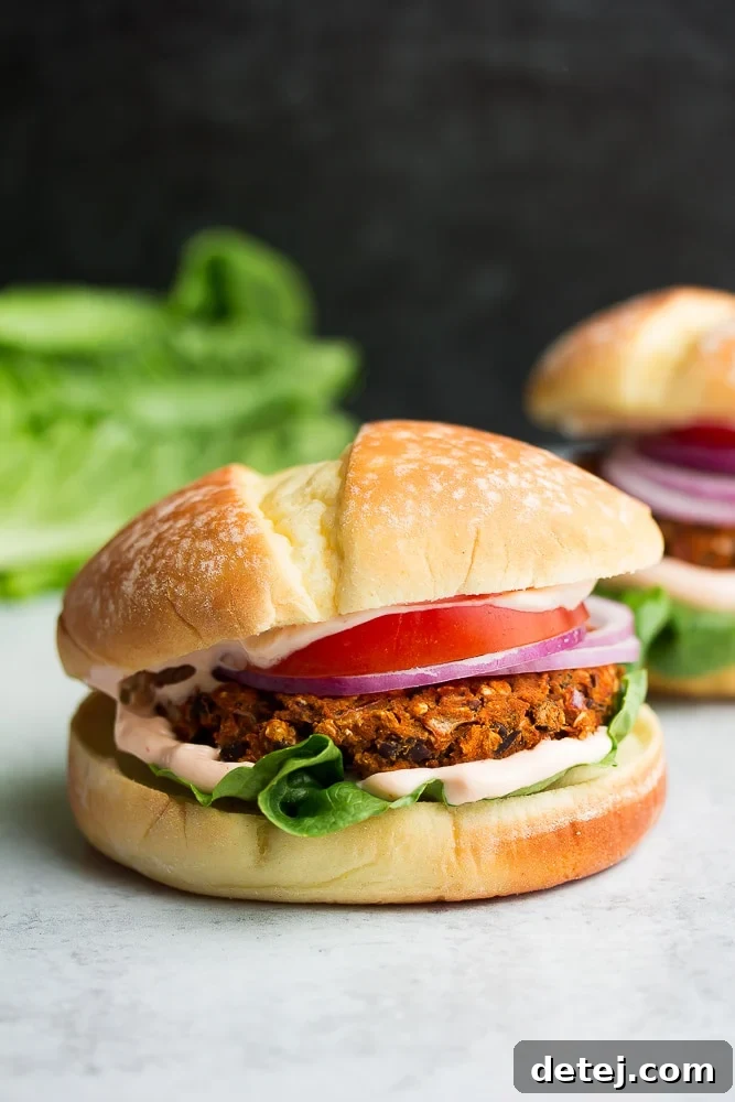 A fully assembled sweet potato black bean burger with an orange and black bean patty, topped with tomato, red onion, Sriracha mayo, and lettuce, all nestled in a bun.