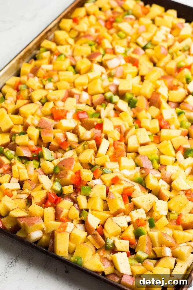 Diced red potatoes, onions, and red and green bell peppers spread in a single, even layer on a large baking sheet, lightly coated with oil and seasonings, ready to be placed in the oven.