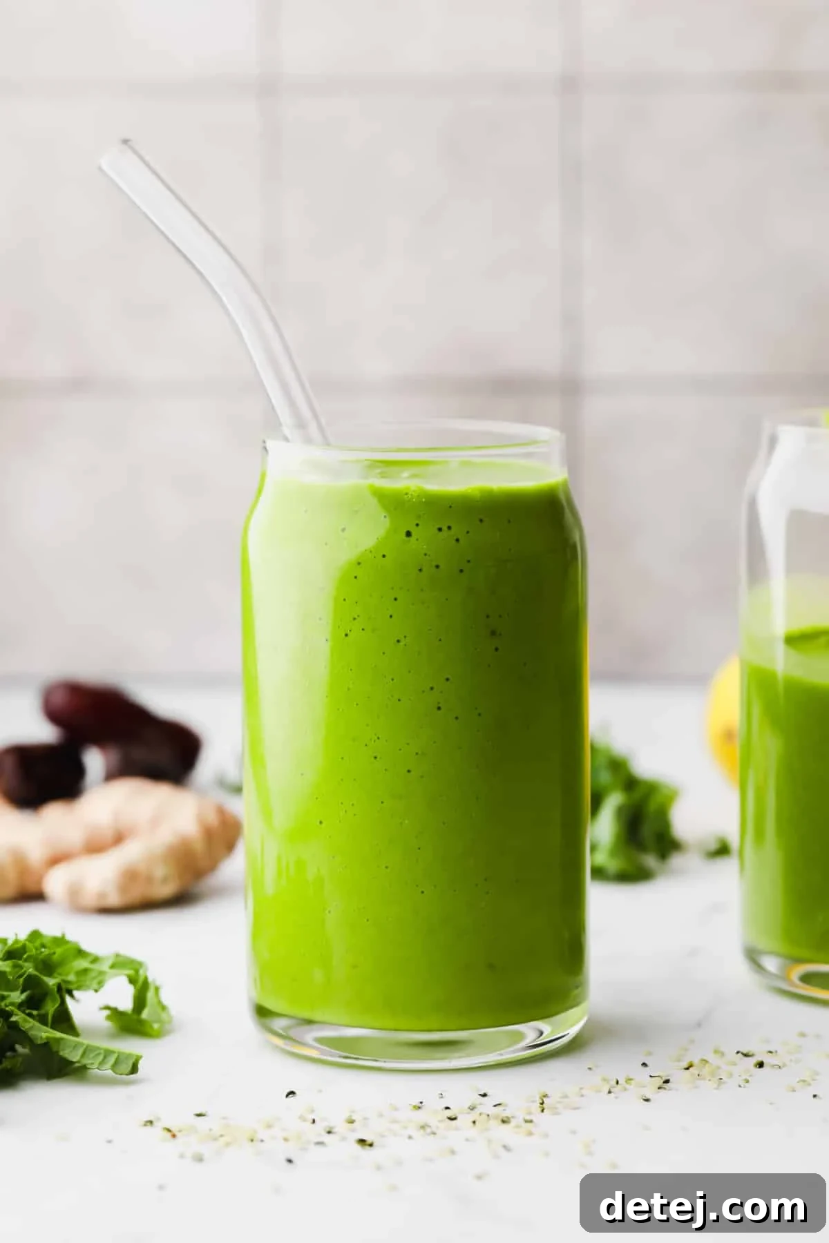 A tall glass cup filled with a smooth, green kale smoothie, complete with a elegant glass straw, set against a modern grey background.