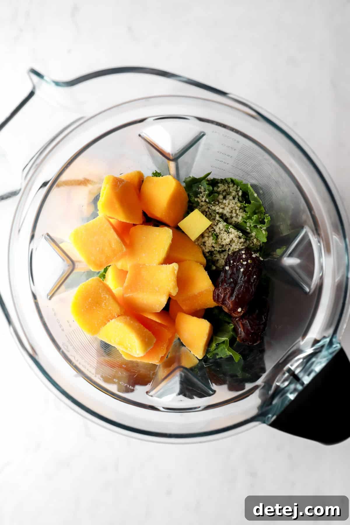Raw ingredients for a kale smoothie, including frozen mango, fresh kale, Medjool dates, and seeds, all perfectly portioned in a blender before blending.