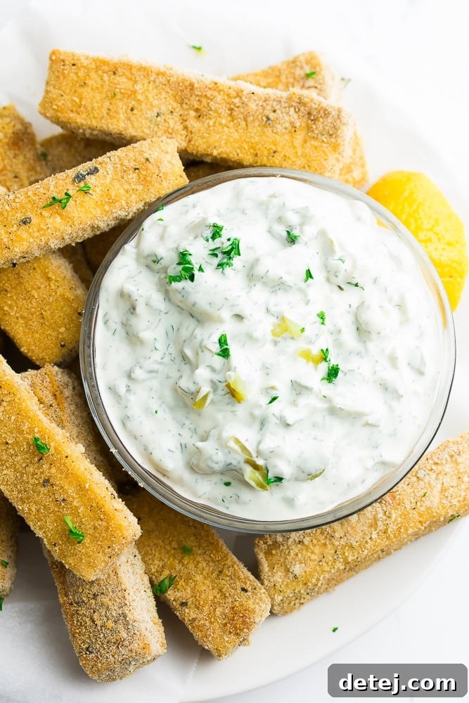 A small, elegant bowl of homemade vegan tartar sauce is beautifully arranged on a rustic wooden board, surrounded by golden-brown, crispy tofu fish pieces, garnished with fresh dill.