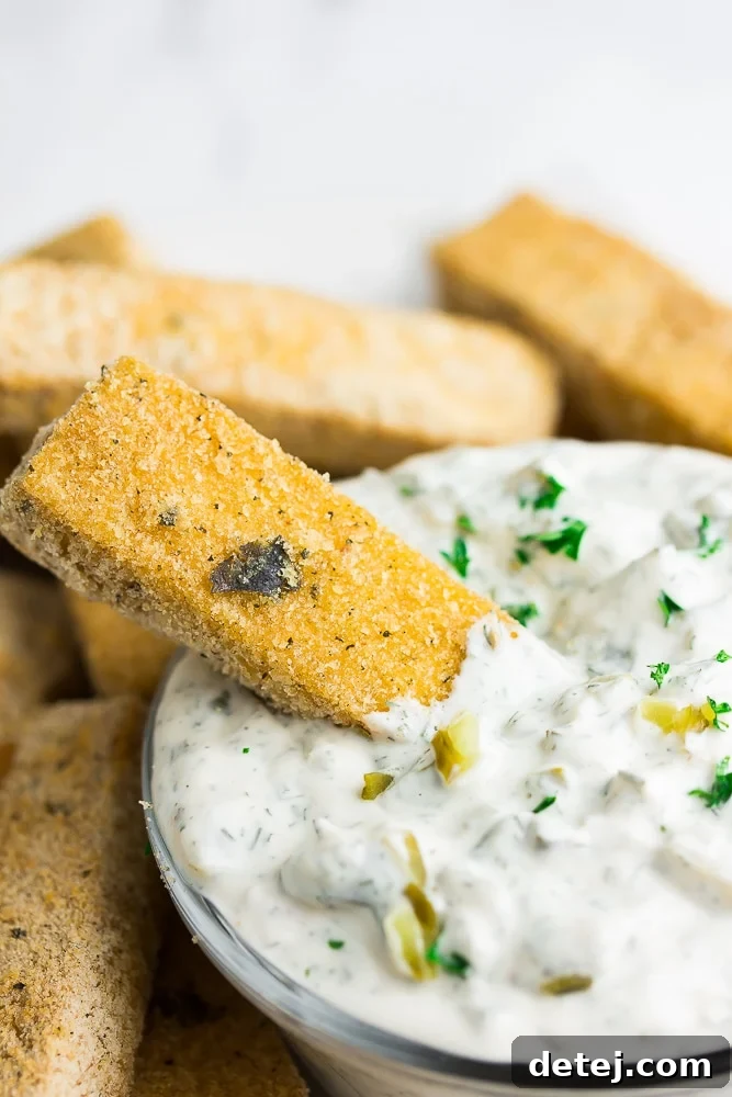 A vegan fish stick being generously dipped into a bowl of creamy white tartar sauce.