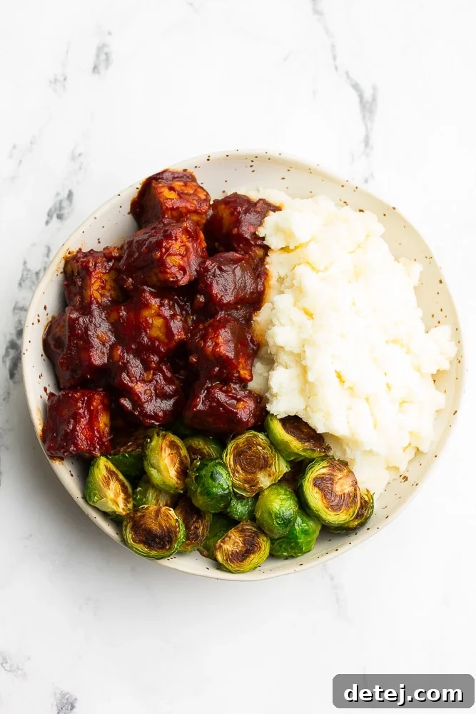 Delicious BBQ Tempeh served with vegan mashed potatoes and roasted brussels sprouts in a white bowl on a white background, highlighting a healthy and quick plant-based meal.