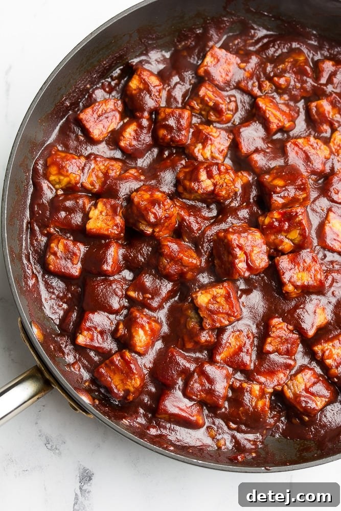 Tempeh cubes generously coated in a rich, dark barbecue sauce within a skillet, simmering to perfection.