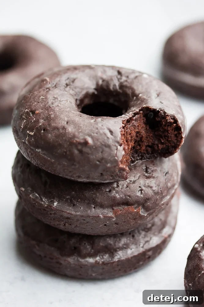 A stack of three decadent vegan chocolate glazed donuts, with a bite taken from the top one to show the soft, moist crumb, all on a simple grey background.
