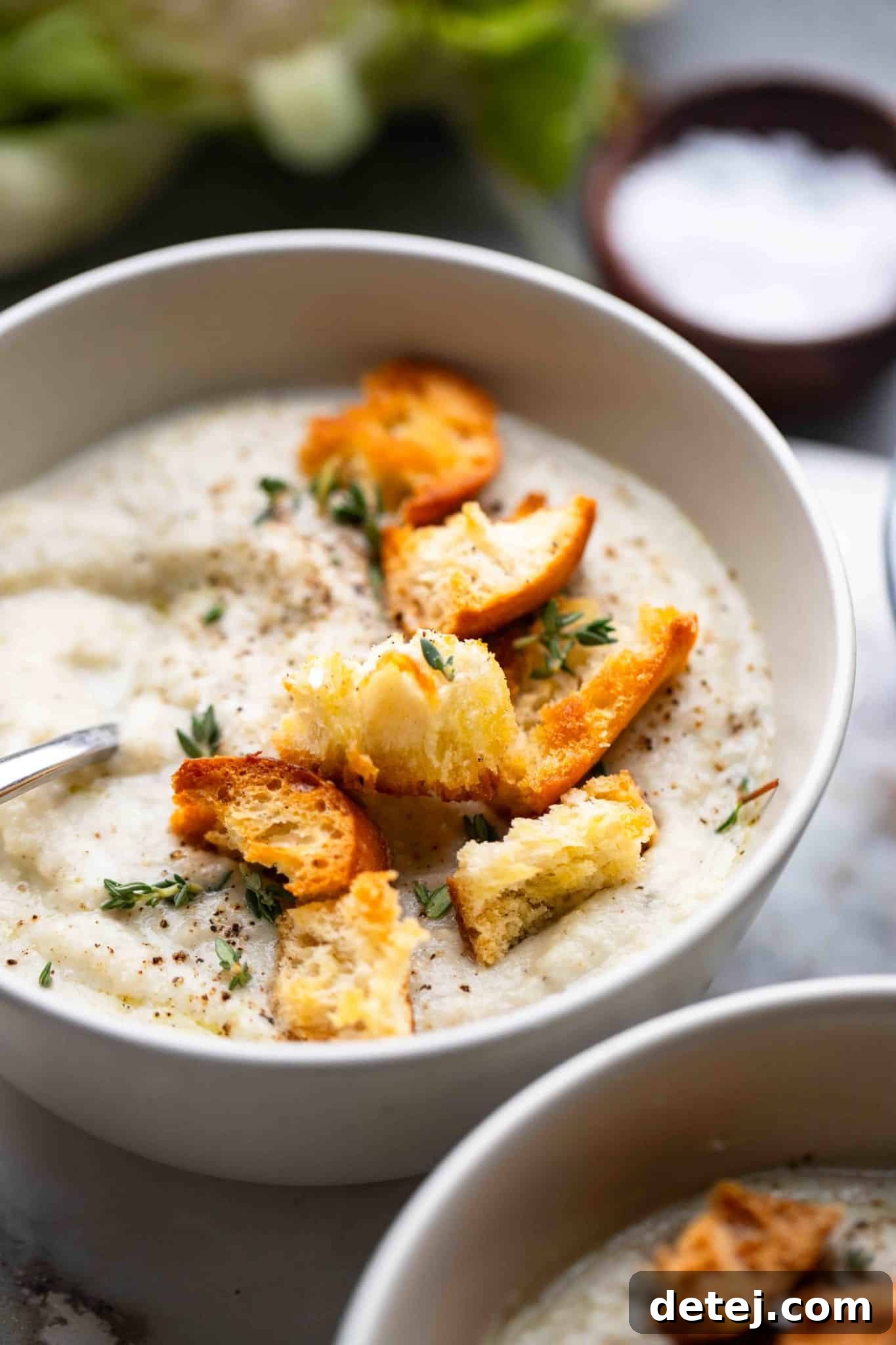 A close-up view of vegan cauliflower soup in a bowl, topped with crunchy croutons and a sprig of fresh rosemary, highlighting its creamy texture.