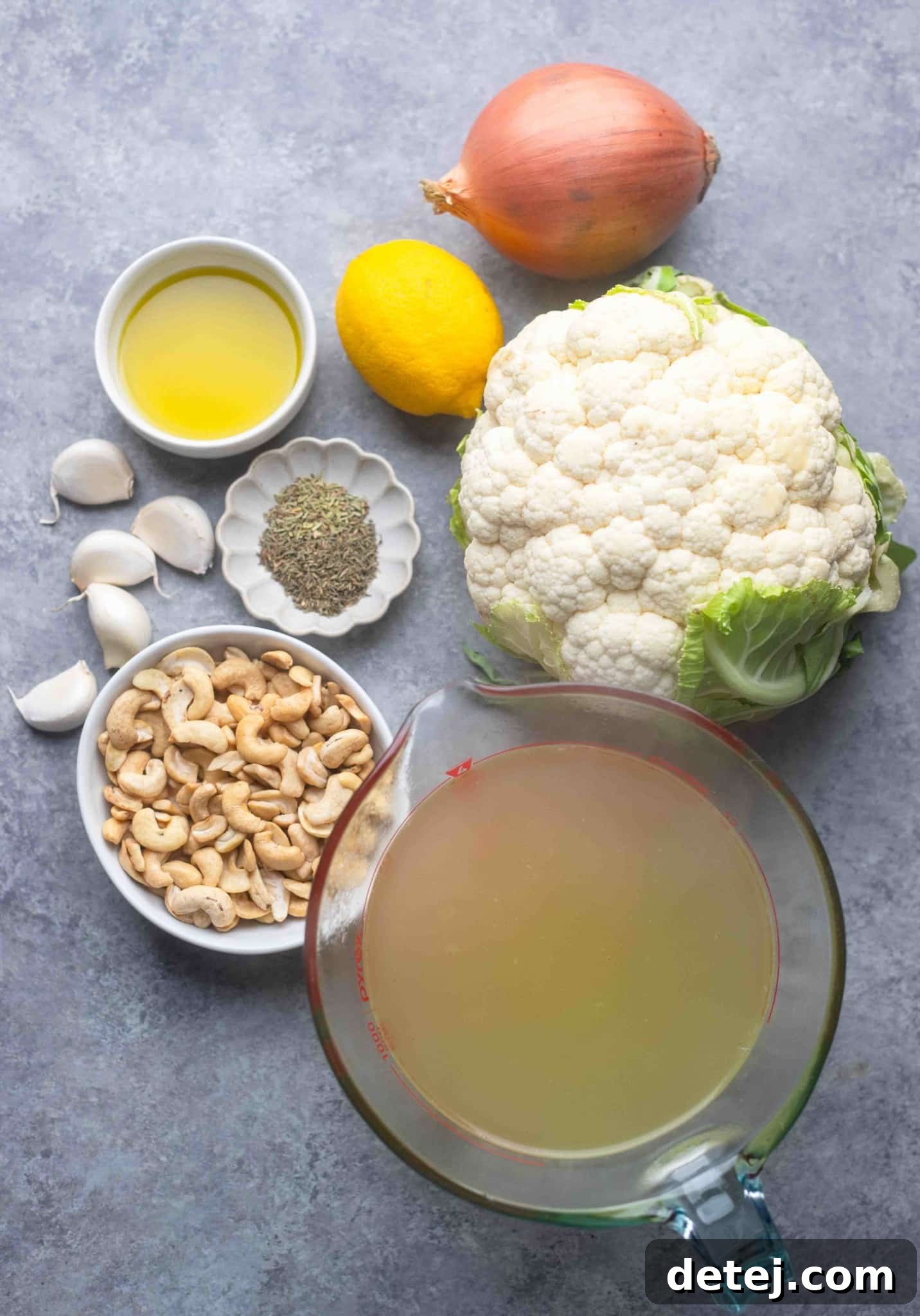 A collection of fresh ingredients for vegan cauliflower soup laid out on a kitchen counter: a head of cauliflower, onions, garlic, cashews, and fresh herbs.