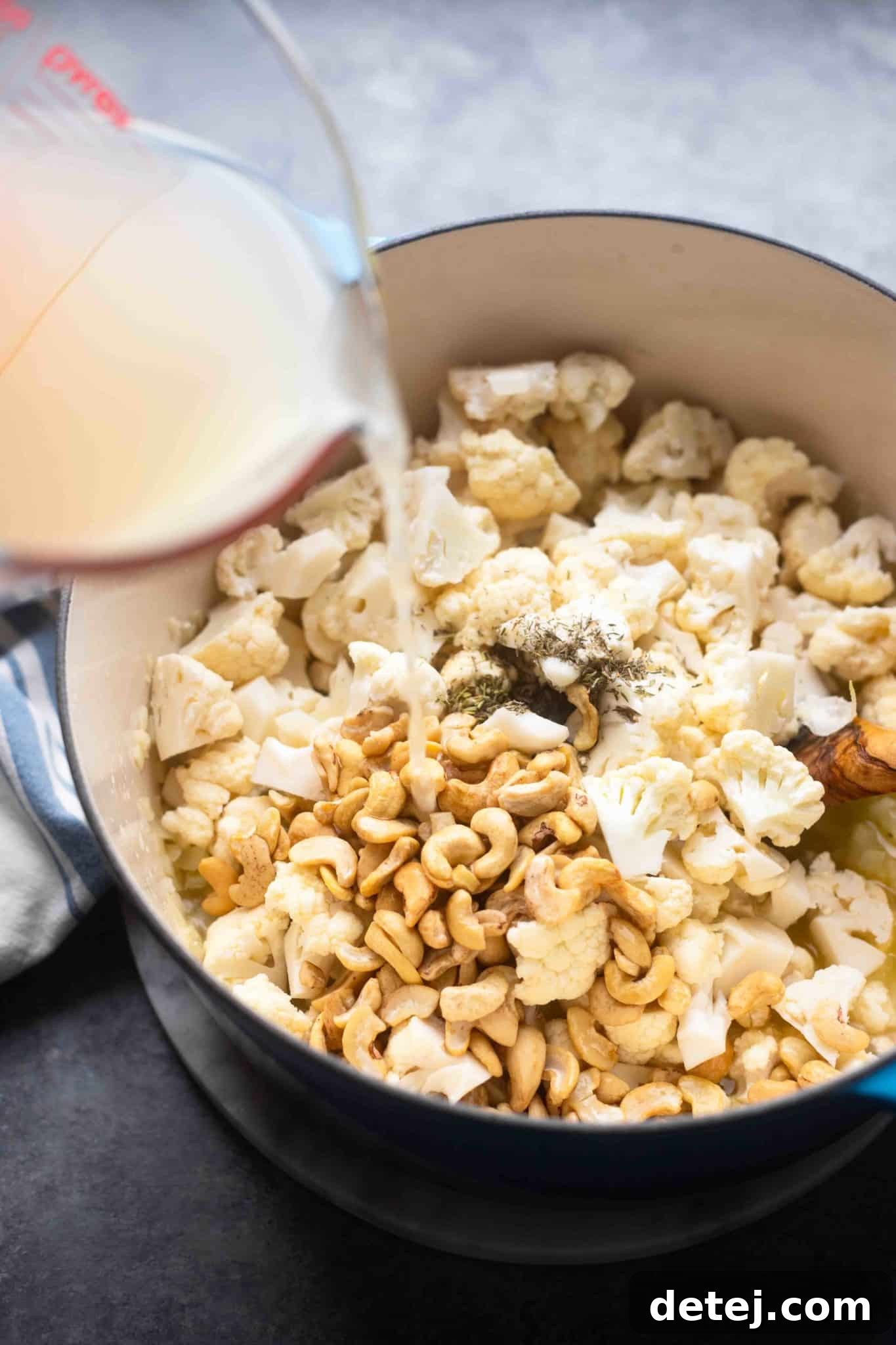 Vegetable broth being poured over cauliflower florets, cashews, and aromatics in a pot, ready for simmering.