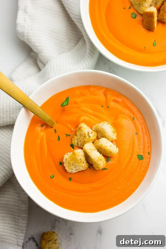 Two bowls of creamy vegan tomato soup with fresh croutons and a golden spoon, set against a clean white background.