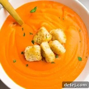 A bowl of creamy vegan tomato soup, garnished with croutons and a golden spoon.