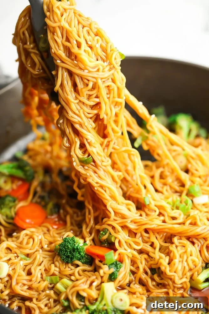 Metal tongs carefully lifting a portion of hot, glistening teriyaki noodles from a large, dark skillet, ready to be served.
