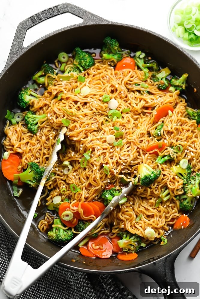 Teriyaki noodles and vibrant vegetables being tossed with metal tongs in a large black skillet, coated in a rich, glossy sauce.