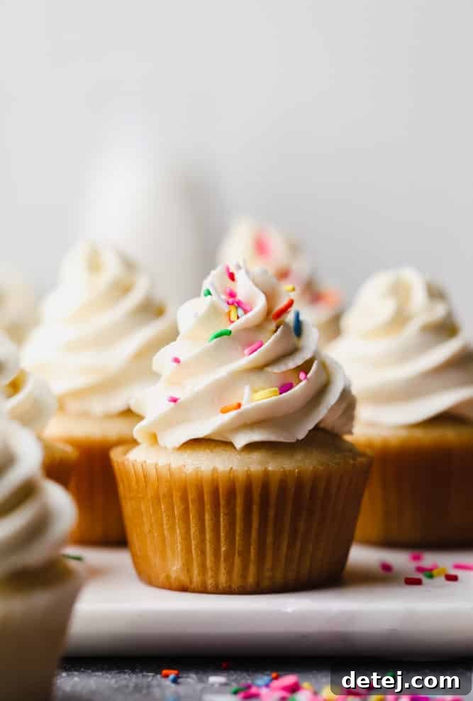 A beautifully frosted vegan vanilla cupcake, perfectly in focus, adorned with colorful sprinkles. More slightly blurred cupcakes are visible in the background, hinting at a delightful batch.