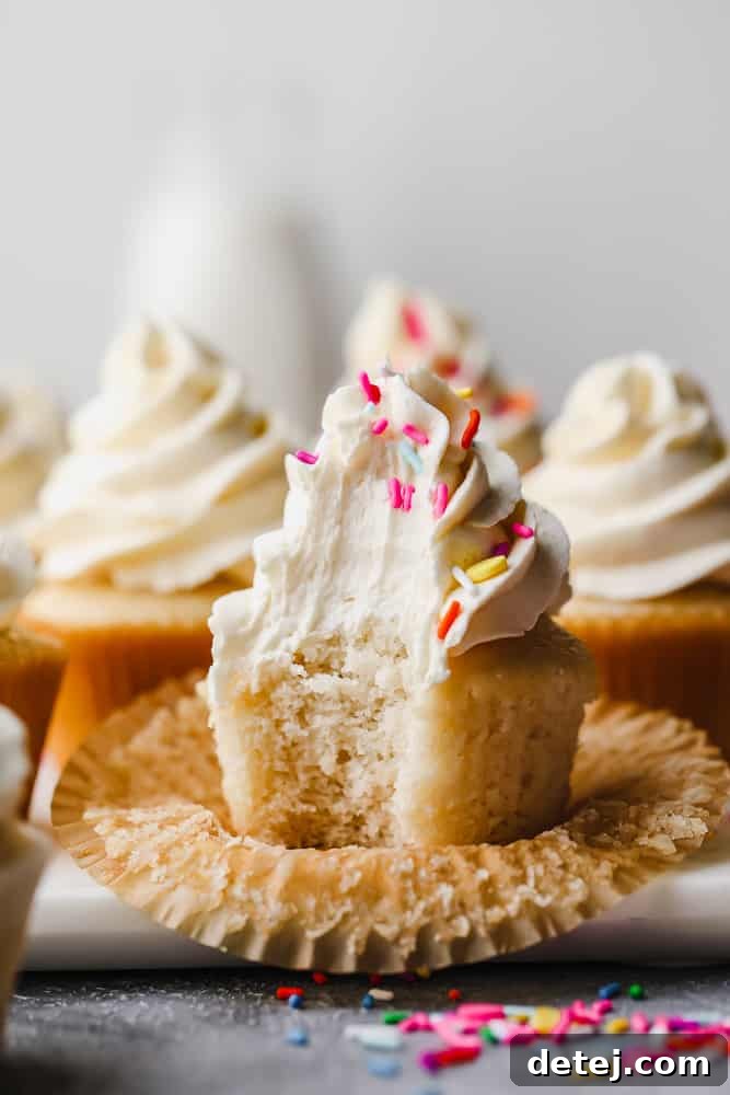 A close-up of a vegan vanilla cupcake with a bite taken out, revealing its moist and fluffy interior, topped with a generous swirl of frosting and colorful sprinkles.