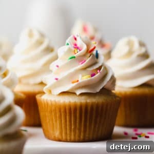 A vibrant display of numerous vegan vanilla cupcakes, adorned with creamy white frosting, some featuring playful sprinkles, arranged for a tempting visual.