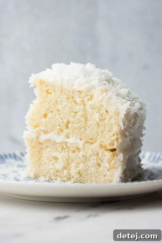 A close-up side view of a perfect slice of vegan coconut cake, showcasing its moist layers, generous creamy frosting, and delicate texture on a clean grey background. A testament to its simple elegance.