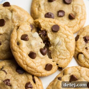 A square photo of a pile of chocolate chip cookies with the top one broken in half, showcasing the gooey chocolate inside.