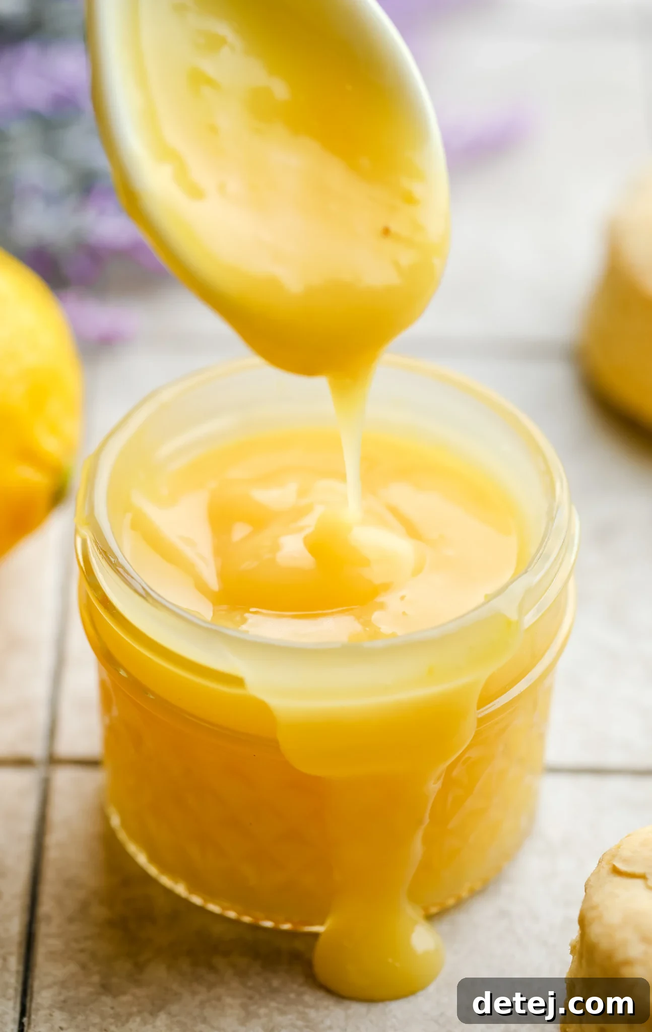 small jar of yellow thickened liquid with a spoon above it on grey background with purple flowers