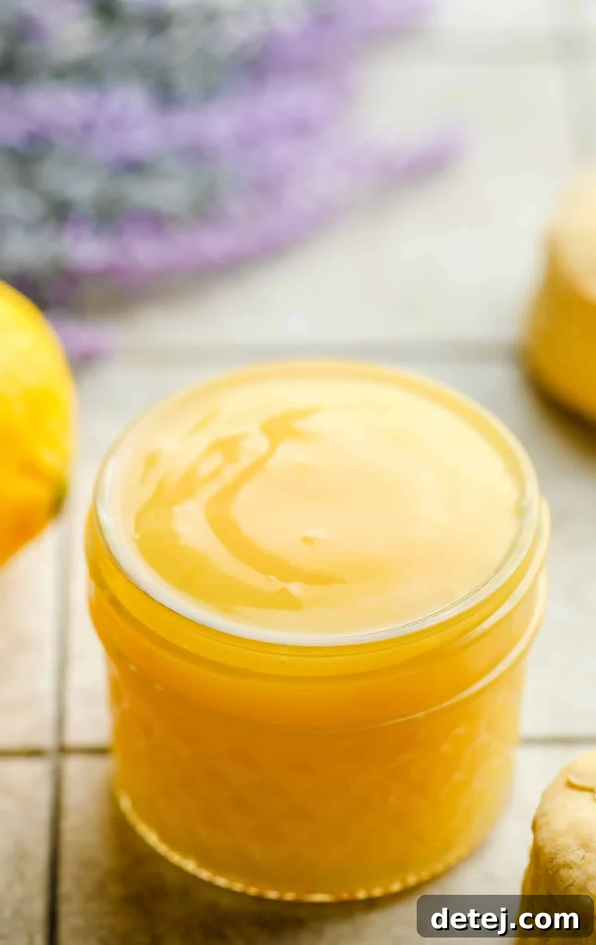 small jar of vegan lemon curd with lavender in background, lemons on a grey squared board