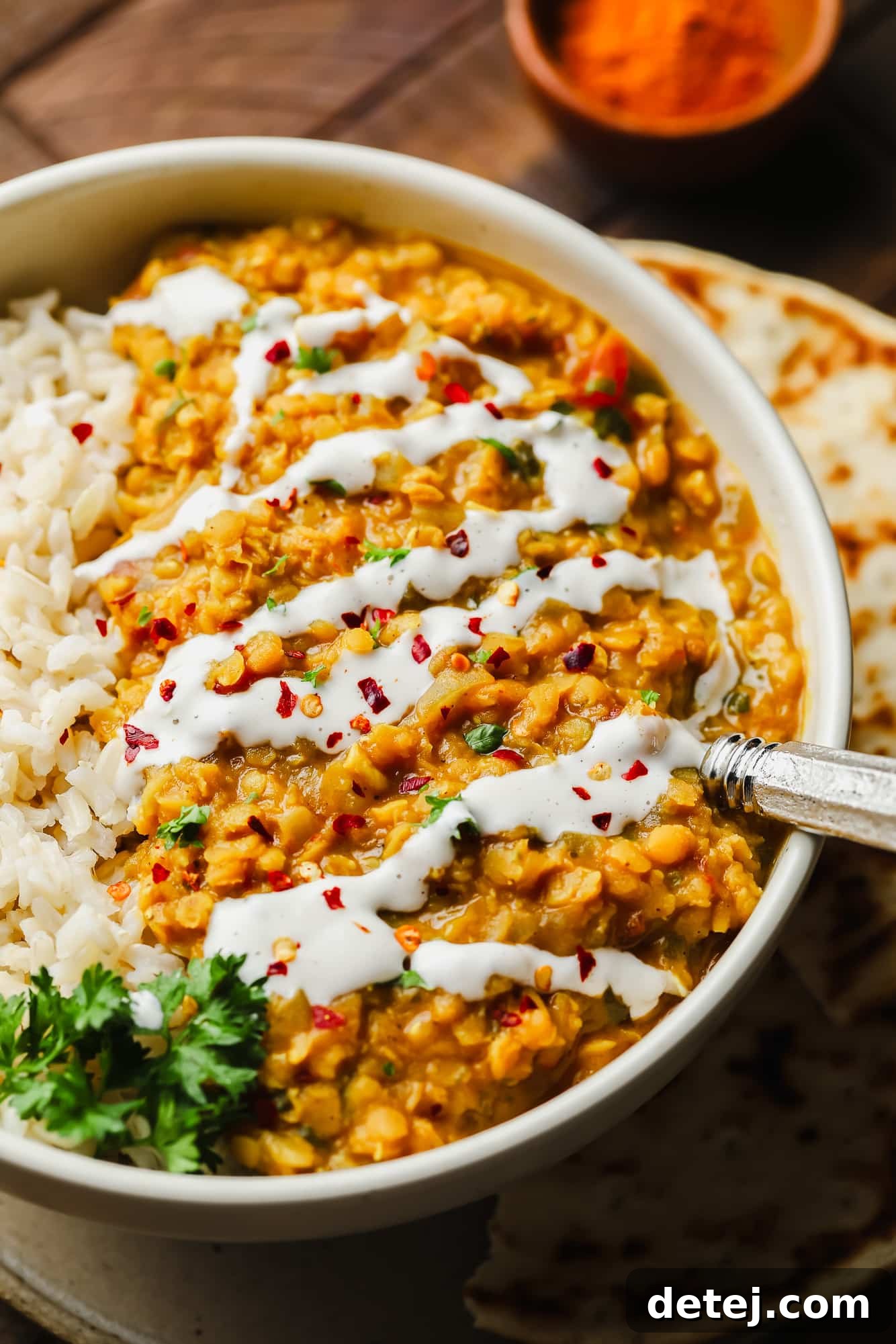 Close up on a bowl of rich red lentil dahl, garnished with a swirl of coconut milk and a sprinkle of red chili flakes.