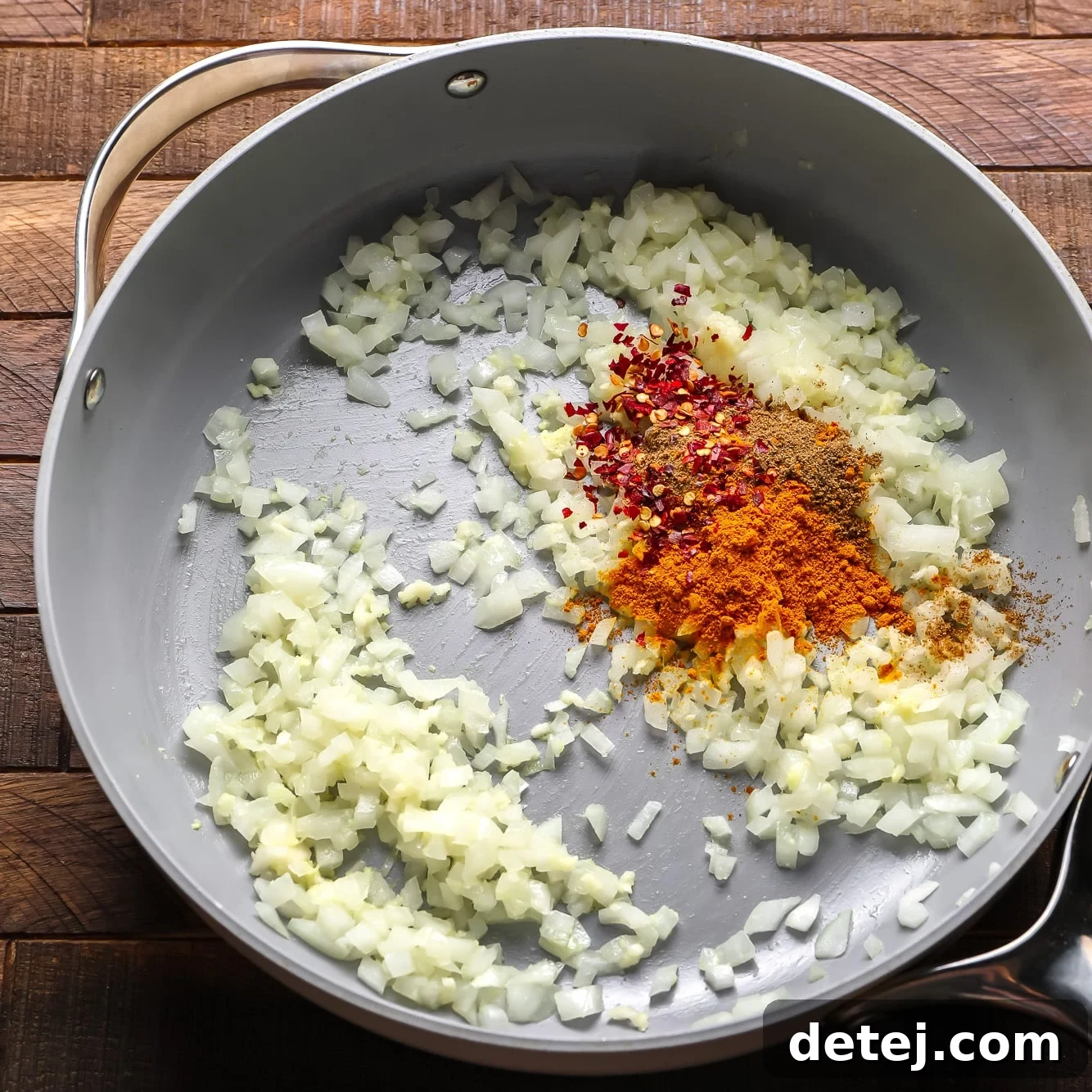 Dry spices, including garam masala and turmeric, sprinkled over sautéed onions in a large grey pan.