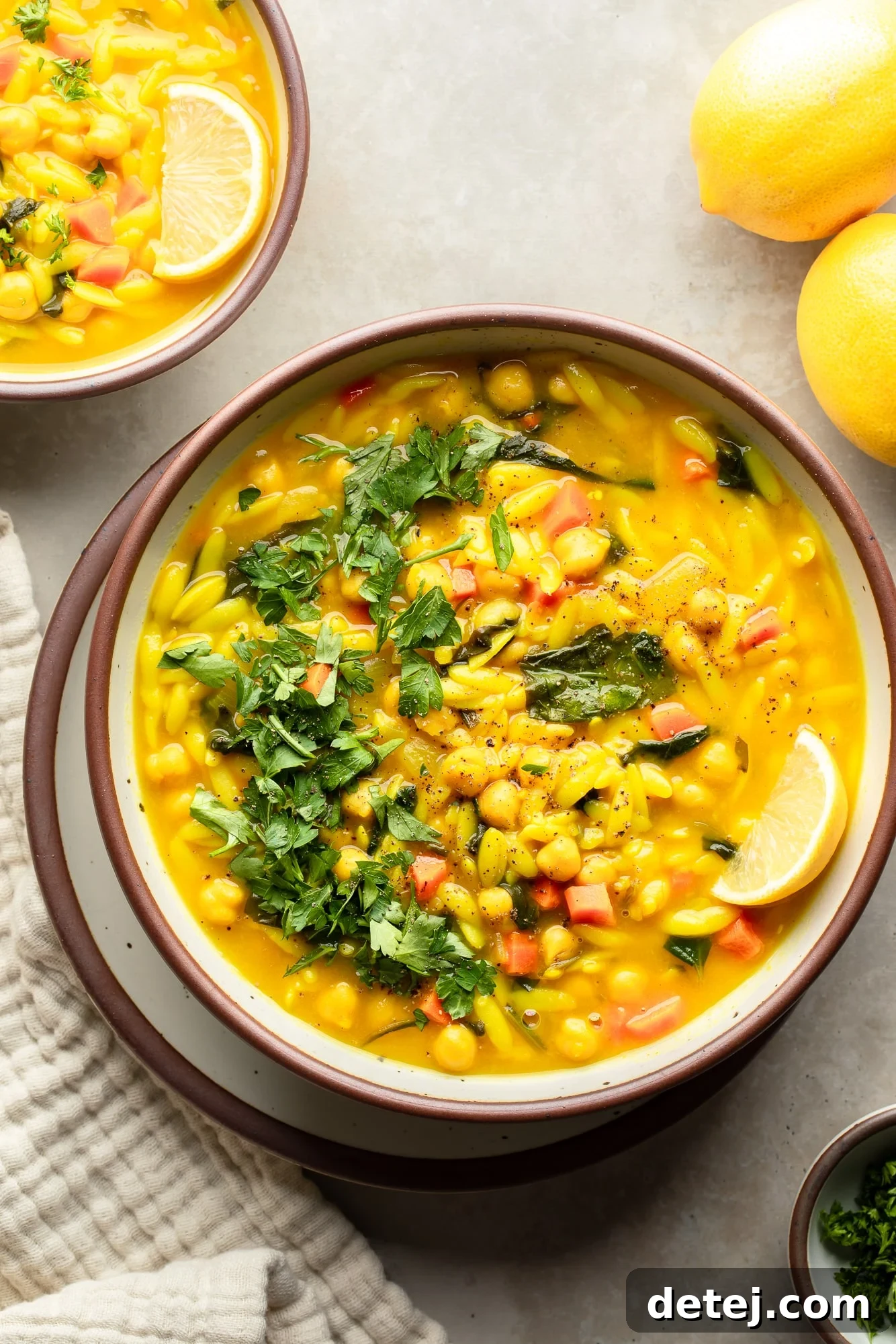 overhead view of a bowl of Golden Orzo Soup with Chickpeas topped with a lemon wedge and green herbs.