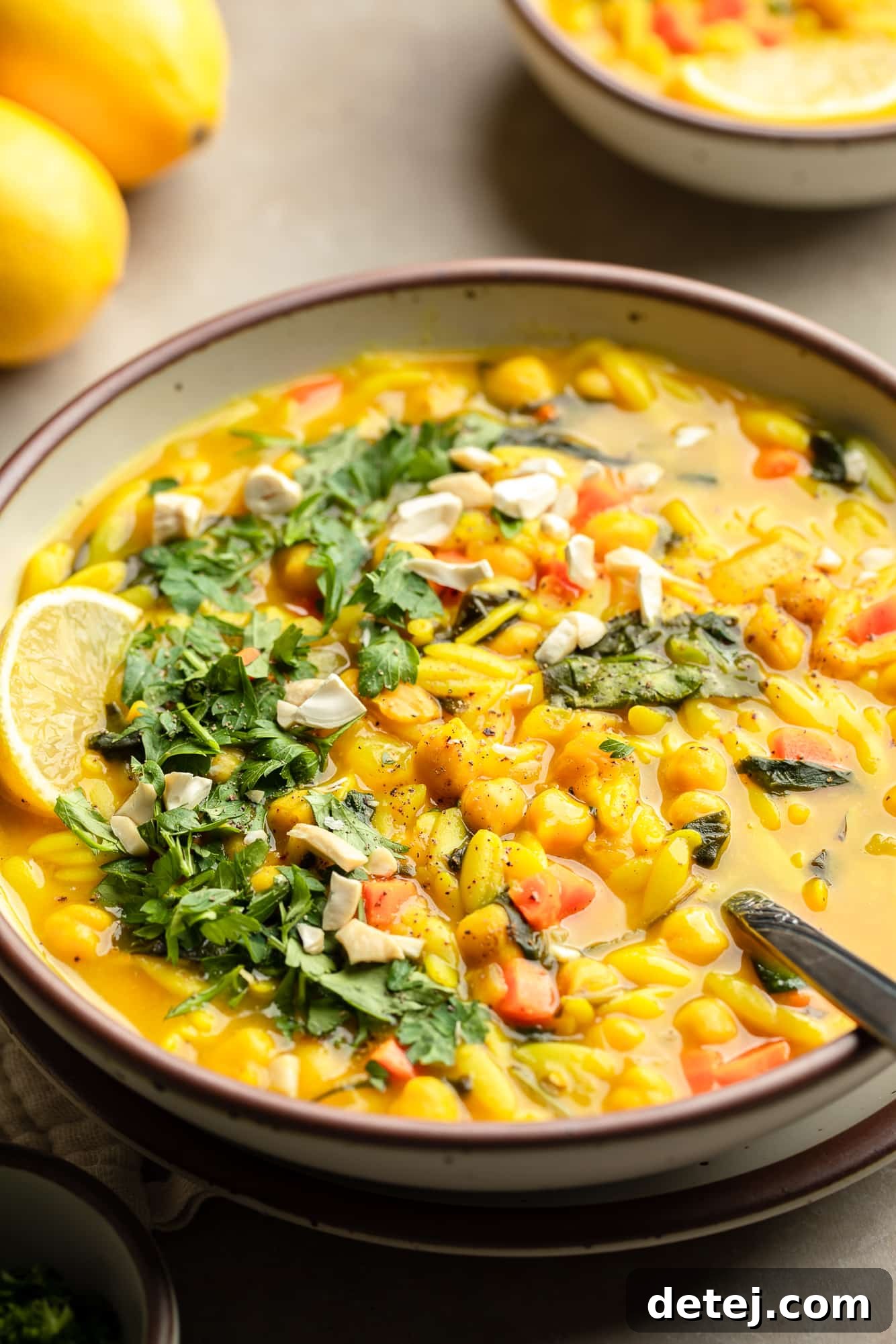 side view of a bowl of Golden Orzo Soup with Chickpeas topped with a lemon wedge, green herbs, and nuts.