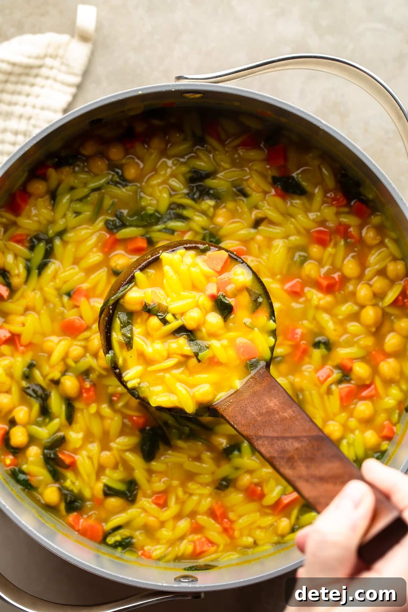 overhead view of a hand lifting a ladle out of a large pot of Golden Orzo Soup with Chickpeas.