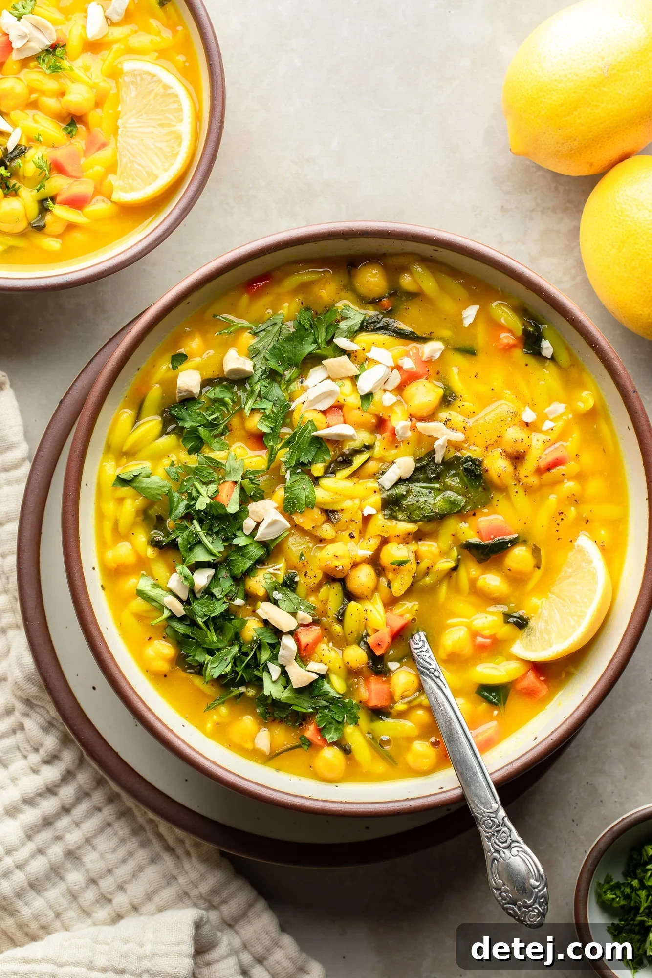 overhead view of a bowl of Golden Orzo Soup with Chickpeas topped with a lemon wedge, green herbs, and nuts.