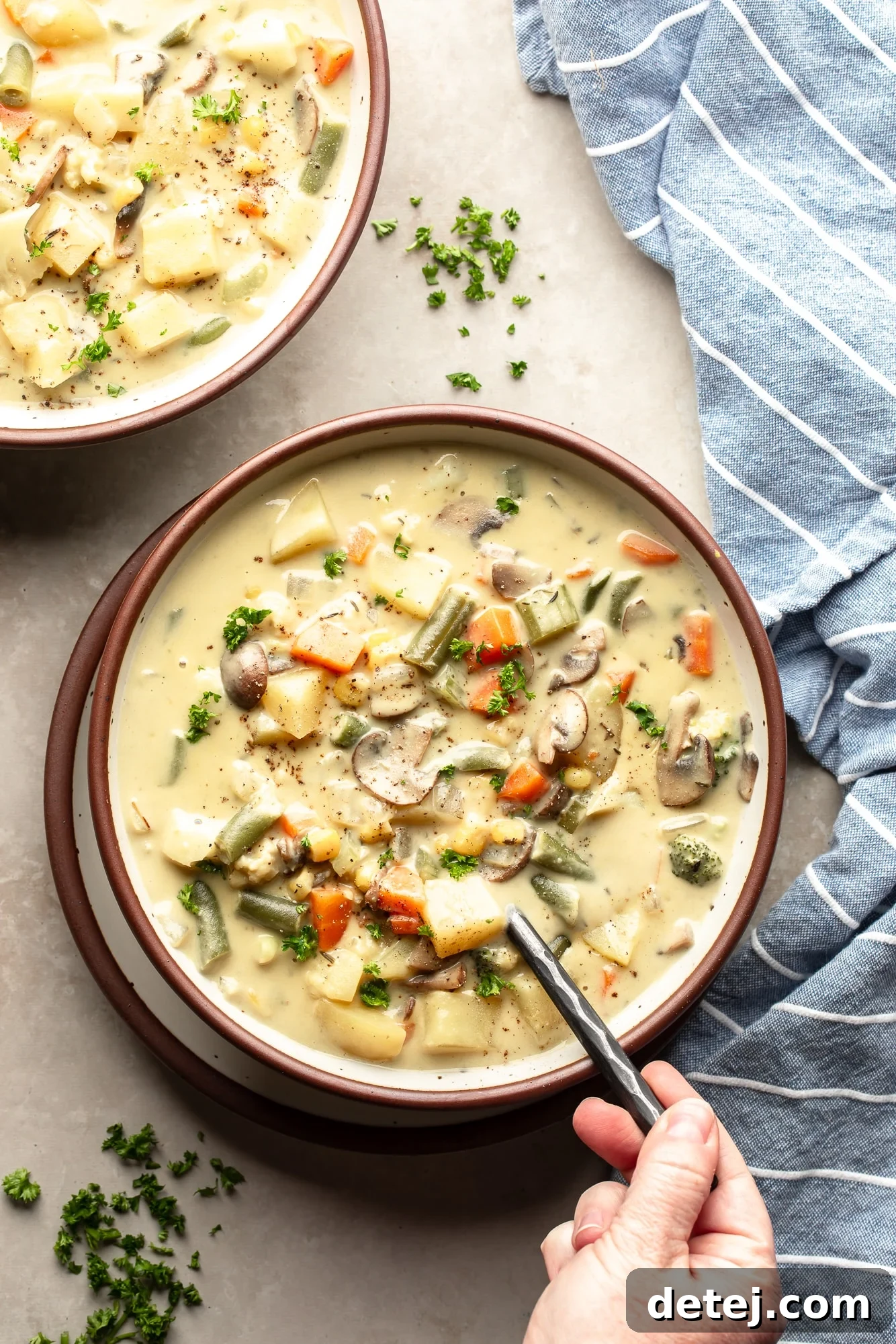 Overhead view of a person's hand holding a spoon in a bowl of Creamy Vegetable Soup, garnished with fresh herbs and a rustic background.