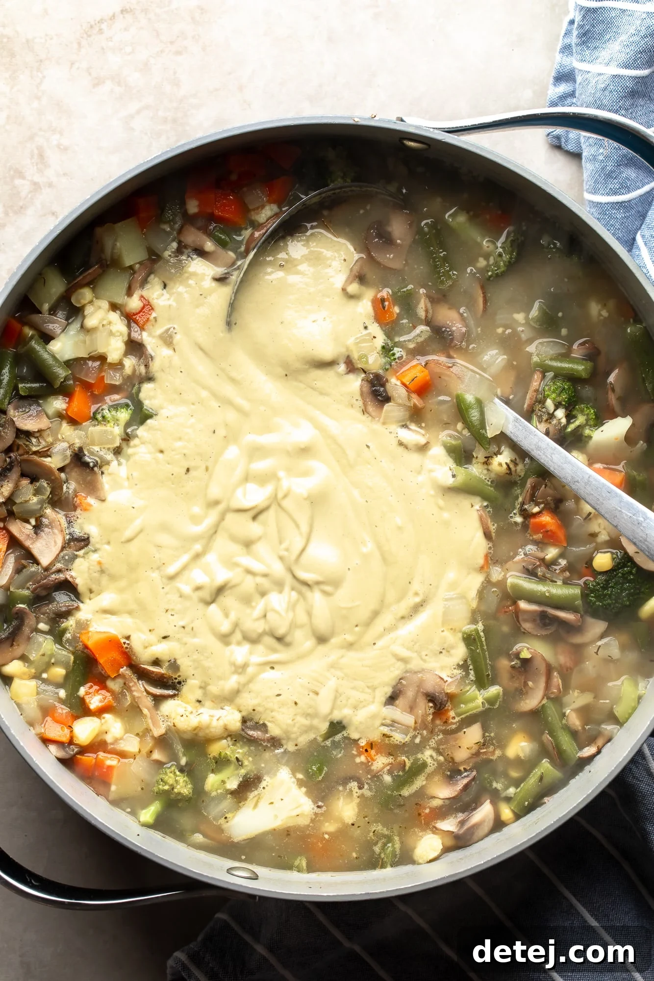Overhead view of a ladle stirring creamy cashew blend into a large pot of Creamy Vegetable Soup, showing the soup thickening and transforming.