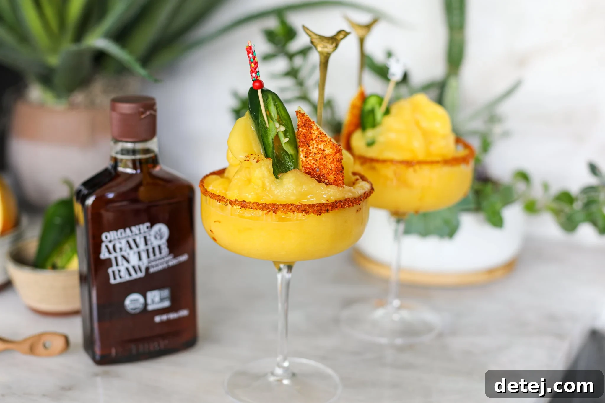 A vibrant Frozen Mango Margarita in a tajin-rimmed glass, garnished with fresh mango and a jalapeno slice, set against a summer backdrop.
