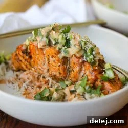Air Fryer Spicy Hasselback Salmon in a bowl with spicy creamy sauce, fresh cucumber, jalapeno, and avocado salad, garnished with sesame seeds and cilantro.